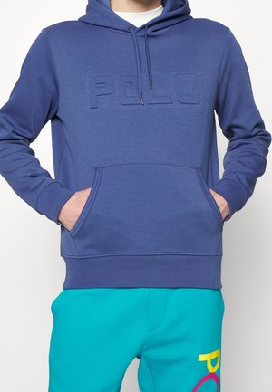 Osoba nosząca niebieską bluzę z kapturem z wytłoczonym napisem "POLO" na piersi oraz turkusowe spodnie z kolorowymi literami "POLO" na udzie.