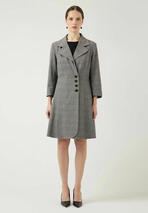 Naine kannab põlveni halli ruudulist mantelkleiti mustade nööpide, 3/4 varrukate, mustade kõrvarõngaste ja mustade teravate kontsadega, seisab frontaalselt.