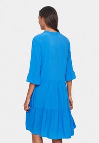 Rochie albastră, în stil A, cu decolteu rotund, mâneci evazate până la trei sferturi și fustă în straturi. Fabricată dintr-un material ușor.