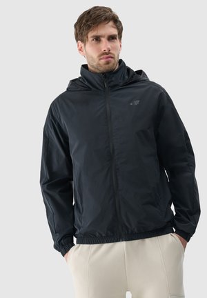 Homme aux cheveux bruns courts portant une veste noire à capuche zippée et un pantalon couleur crème, debout les mains dans les poches devant un fond clair.
