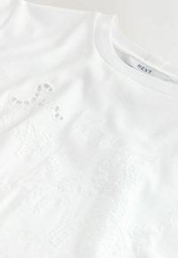 Tessuto bianco con un delicato motivo tropicale in rilievo che include palme, stelle marine e forme di girasole, e una scollatura rotonda con etichetta NEXT.