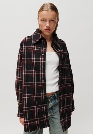 Jeune personne aux cheveux plaqués en arrière portant une chemise à carreaux noire et rouge boutonnée par-dessus un débardeur blanc et un jean bleu clair.