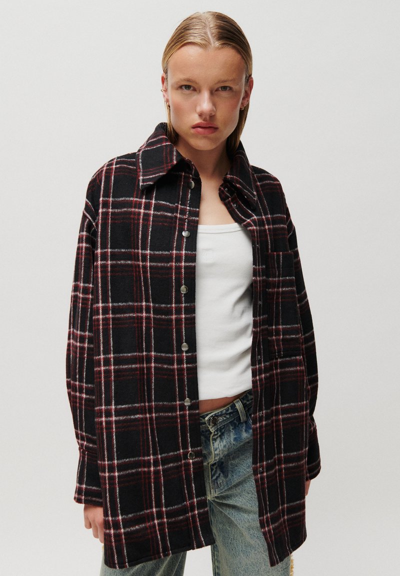 Jeune personne aux cheveux plaqués en arrière portant une chemise à carreaux noire et rouge boutonnée par-dessus un débardeur blanc et un jean bleu clair.