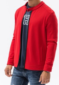 Giacca rossa con zip e collo alto, dotata di tasche laterali e design aderente, indossata sopra una camicia blu navy con stampa di lettere bianche.