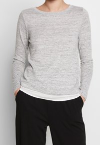 Suéter de manga larga gris claro con un patrón de punto texturizado y un cuello redondo. Presenta un bajo de canalé blanco y se lleva sobre pantalones negros.