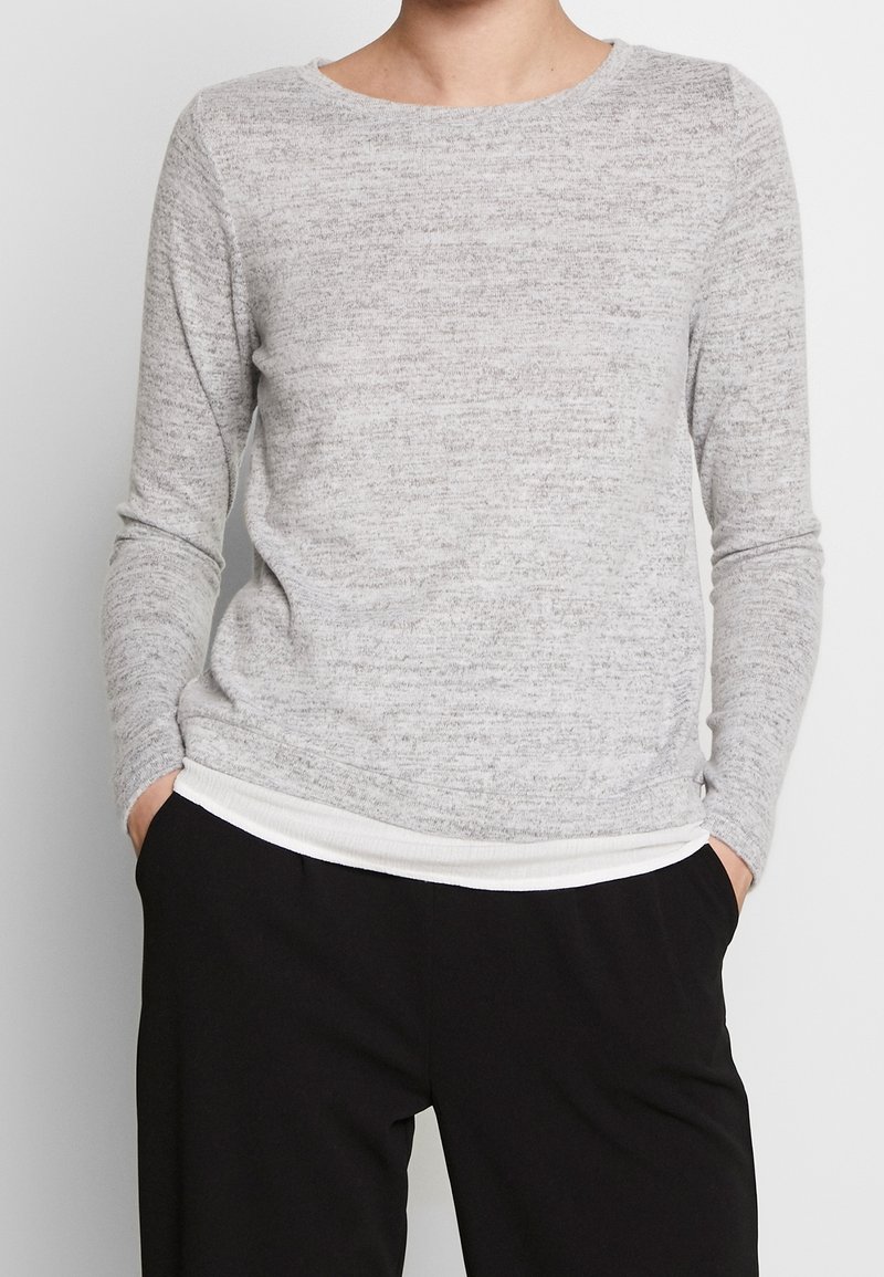 Suéter de manga larga gris claro con un patrón de punto texturizado y un cuello redondo. Presenta un bajo de canalé blanco y se lleva sobre pantalones negros.