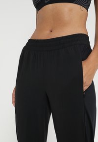 Černé sportovní kalhoty s přiléhavým pasem, vyrobené z lehkého materiálu. Obsahují boční kapsy a hladkou texturu, navrženy pro pohodlí.