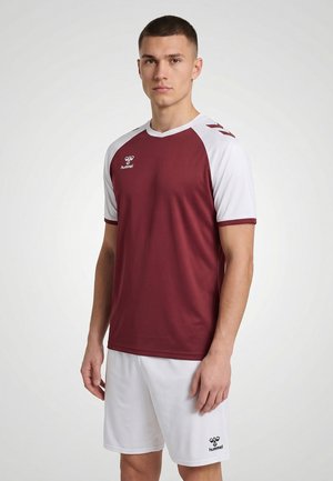 Junger Mann stehend, trägt ein burgunderrotes und weißes Sporttrikot sowie weiße Shorts mit Hummel-Logo, vor einem schlichten hellen Hintergrund.