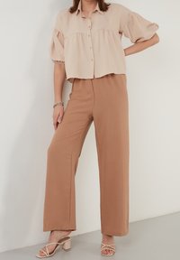 Beżowa krótka koszula zapinana na guziki z bufkami, w połączeniu z jasnobrązowymi spodniami o szerokich nogawkach. Miękka tkanina, luźny krój, subtelna tekstura i prosty design.