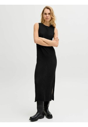 Modelo femenina de pie con los brazos cruzados, vistiendo un vestido midi de punto negro sin mangas y botas negras hasta la rodilla sobre un fondo liso.