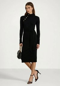 Mulher vestindo um vestido midi preto justo de manga longa com botões dourados no ombro, segurando uma bolsa preta e usando saltos pretos.