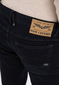 Jean en denim foncé avec un tissu texturé, présentant une étiquette visible avec le nom de la marque, le logo et des détails de couture. Deux poches arrière incluses.