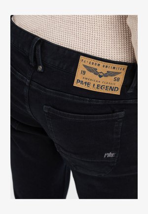 Tamni traperici s teksturiranim materijalom, s vidljivom etiketom koja sadrži naziv brenda, logotip i detalje šivanja. Uključena su dva stražnja džepa.