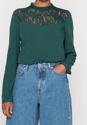 Vrouw draagt een donkergroen blouse met lange mouwen en kanten inzetstuk bij de hals, gecombineerd met een hoog uitgesneden blauwe spijkerbroek, handen in de zakken, voor een effen achtergrond.