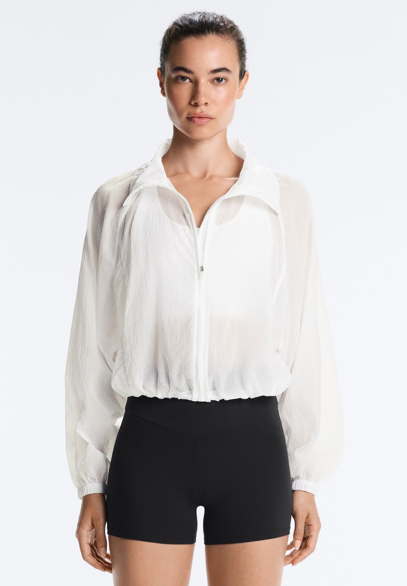Femme aux cheveux foncés attachés, portant une veste légère blanche zippée et un short noir taille haute ajusté, debout devant un fond uni.