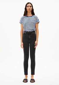 Camiseta a rayas azul y blanca con mangas enrolladas, jeans skinny negros de cintura alta y sandalias oscuras sin hebilla, de pie contra un fondo blanco.