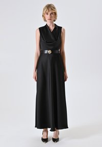 Donna con capelli biondi corti che indossa un maxi abito nero senza maniche con scollatura drappeggiata, cintura nera con fibbia a forma di fiore dorata e tacchi neri.