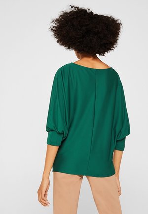 Femme aux cheveux bouclés portant un haut ample vert émeraude à manches trois-quarts et un pantalon beige, vue de dos sur un fond uni.