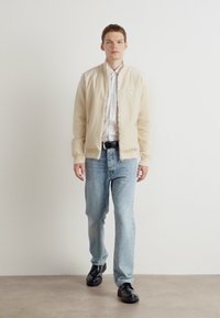 Beige bomberjacka med ribbade ärmar, bärs över en vit skjorta. Ljusblå jeans och svarta skor fulländar outfiten. Modellen står framför en enkel bakgrund.