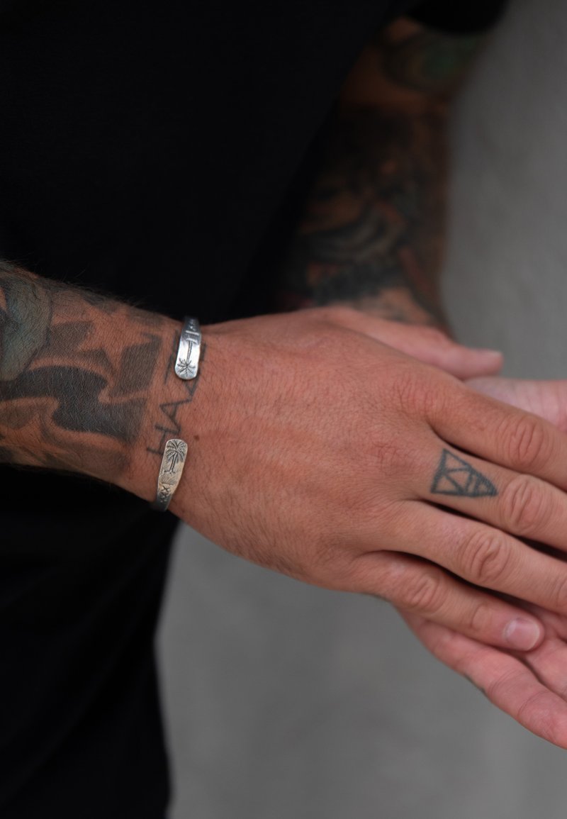 Sidabriniai apyrankės ant tatuiruotėmis padengtos rankos. Viena apyrankė turi palmės medžio dizainą, kita yra su lygiais, tiesiais kraštais. Oda yra vidutinės spalvos.