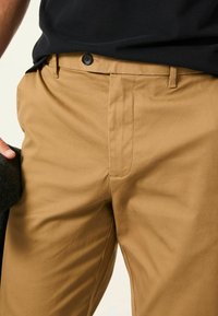 Beige Baumwollhose mit glatter Textur, ausgestattet mit einem Knopfverschluss vorne und seitlichen Taschen. In der linken Hand wird ein schwarzer Stoff gehalten.