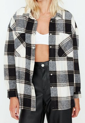 Kvinde iført en åben sort, hvid og grå ternet skjorte over en hvid croptop og højtaljede sorte læderbukser.