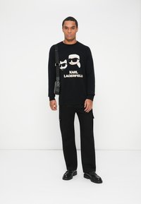 Fekete kötött pulóver, napszemüveggel és a "KARL LAGERFELD" szöveggel ellátott grafikai dizájnnal, fekete cargo nadrággal és fekete cipővel párosítva.