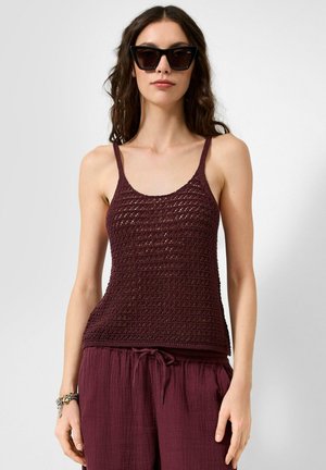 Vrouw met donkere zonnebril, een mouwloze gestructureerde bordeauxrode top en losse bordeauxrode broek met koordsluiting in de taille tegen een effen achtergrond.