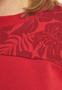 Rotes Sportshirt mit einem strukturierten Blumenmuster auf den Schultern, das ein kleines gesticktes Logo in passendem roten Faden aufweist.