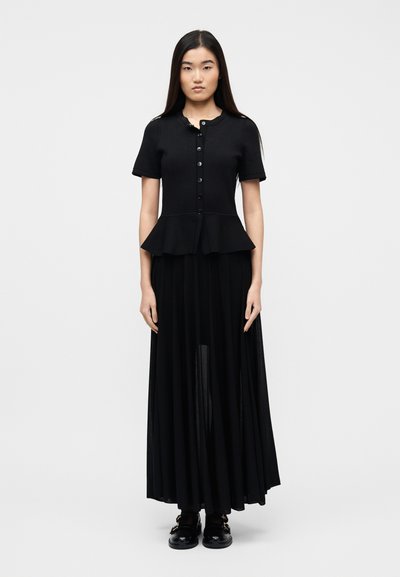 Femme aux cheveux noirs raides portant un haut péplum noir à manches courtes et une longue jupe noire, debout devant un fond clair uni.