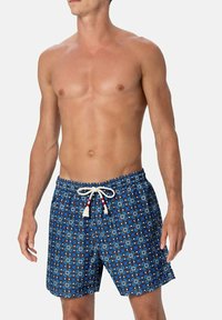 Uomo a torso nudo che indossa pantaloni da bagno blu a fantasia con un cordoncino bianco, in piedi contro uno sfondo chiaro e uniforme.