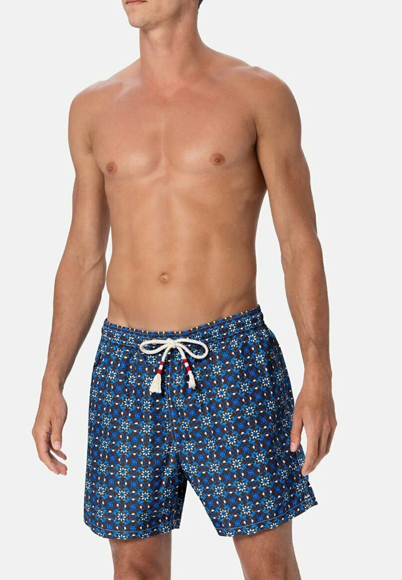 Uomo a torso nudo che indossa pantaloni da bagno blu a fantasia con un cordoncino bianco, in piedi contro uno sfondo chiaro e uniforme.