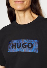Černé bavlněné tričko s modro-černým hadím vzorem a slovem "HUGO" v tučném černém písmu umístěným na hrudi.