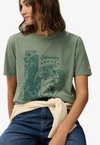 Olivijnkleurig T-shirt met een grafische print van een vogel en een sierlijk ontwerp. Korte mouwen, ronde hals, katoenen materiaal en een losse pasvorm.