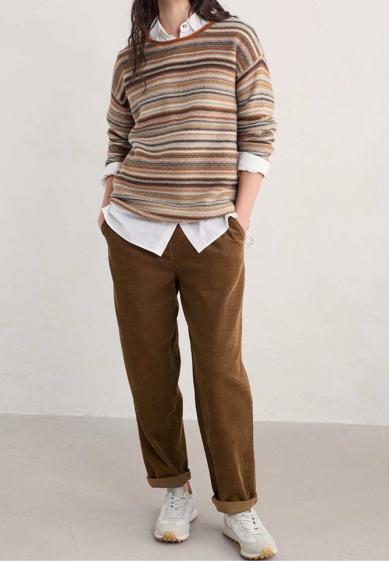 Personne portant un pull rayé marron, beige et orange par-dessus une chemise blanche, un pantalon en velours côtelé marron et des baskets blanches, debout les mains dans les poches.