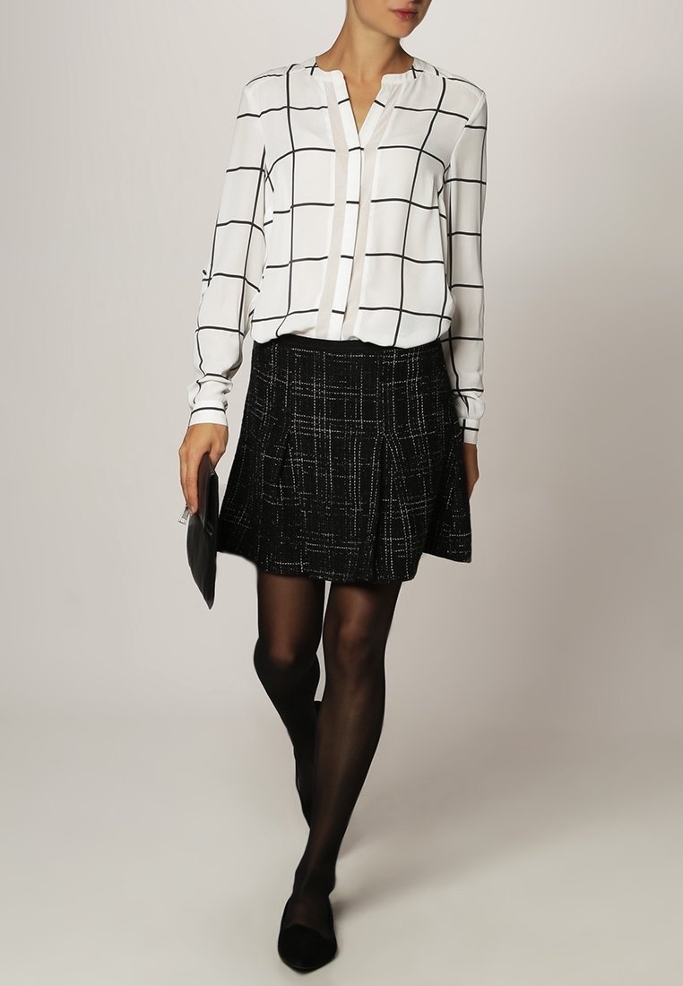 Blusa bianca con motivo a griglia nera, abbinata a una gonna nera testurizzata. Il look completo include calze nere e scarpe basse.