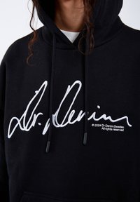 Black hoodie featuring a large white embroidered signature logo. Drawstrings with black aglets and a front pocket detail. Soft fabric texture.