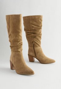 Knee-high beige mokkasini-saappaat, joissa on rypytetty muotoilu, teräväkärkiset ja tukevat korko. Varustettu sivuvetoketjulla helppoon pukemiseen.