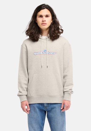 Jong persoon met lang donker haar die een beige Quiksilver hoodie en blauwe spijkerbroek draagt, staand tegen een effen achtergrond.