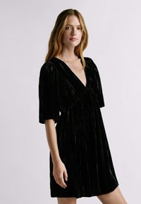 Robe en velours noir avec un décolleté en V profond, des manches courtes bouffantes et une taille froncée. Présente une texture verticale et une coupe au genou.