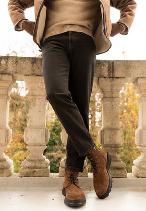Personne portant des bottes en daim marron, un pantalon noir et une veste beige, debout jambes croisées sur un balcon en pierre à l'extérieur en automne.