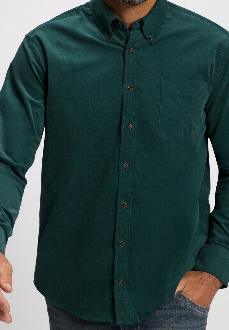 Mörkgrön kordflöjelsskjorta med krage som knäpps, bröstficka och bruna knappar; har en mjuk textur och rundad nederkant.