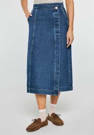 Kobieta w niebieskiej spódnicy dżinsowej o długości do połowy łydki z zapięciem na guziki z przodu, białe skarpetki i brązowe zamszowe buty na sznurowadła na jednolitym tle.