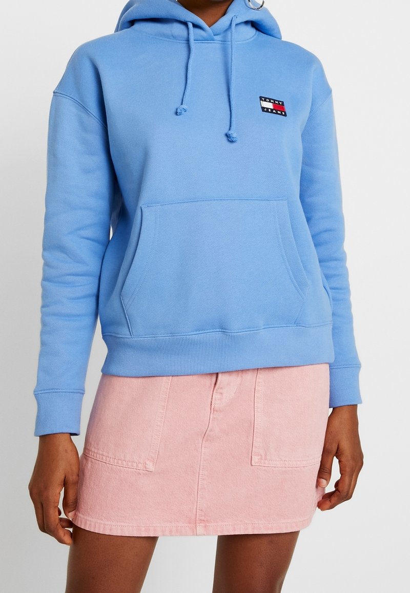 Felpa con cappuccio blu chiaro realizzata in tessuto morbido, con tasca frontale e piccolo patch con logo. Abbinata a una gonna di denim rosa.