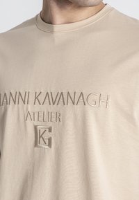 Béžové bavlnené tričko s reliéfnym textom "GIANNI KAVANAGH ATELIER" a logovým akcentom, okrúhlym výstrihom a hladkou textúrou.