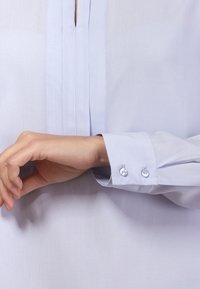 Jasnoniebieska koszula z guzikami z plisowanym przodem i mankietem z dwoma guzikami. Tkanina wydaje się gładka i ma miękką teksturę.