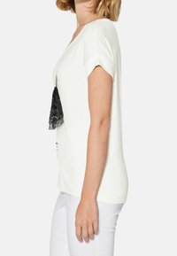 Camiseta blanca de manga corta, con un detalle de encaje negro en el frente. Tejido suave con un corte relajado, mostrada de lado.