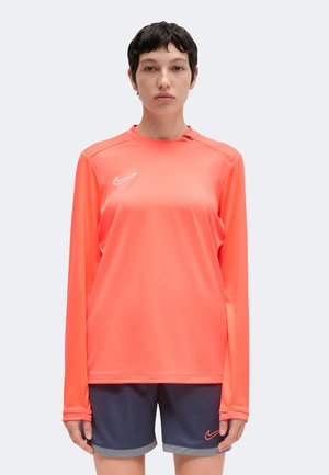 Personne aux cheveux courts et noirs portant un maillot de sport à manches longues couleur corail vif et un short bleu foncé avec des logos Nike, debout devant un fond uni.