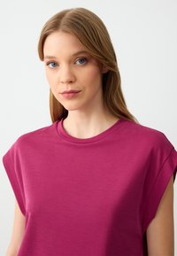 Top magenta a maniche corte con una texture liscia, scollo rotondo e polsini arrotolati, caratterizzato da una vestibilità ampia e un tessuto leggero.