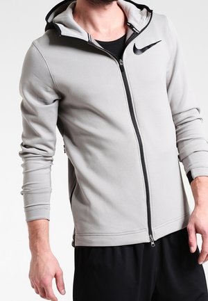 Mand iført lys grå Nike hættetrøje med lynlås og sort logo samt sorte bukser, beskåret ved halsen og midt på låret.